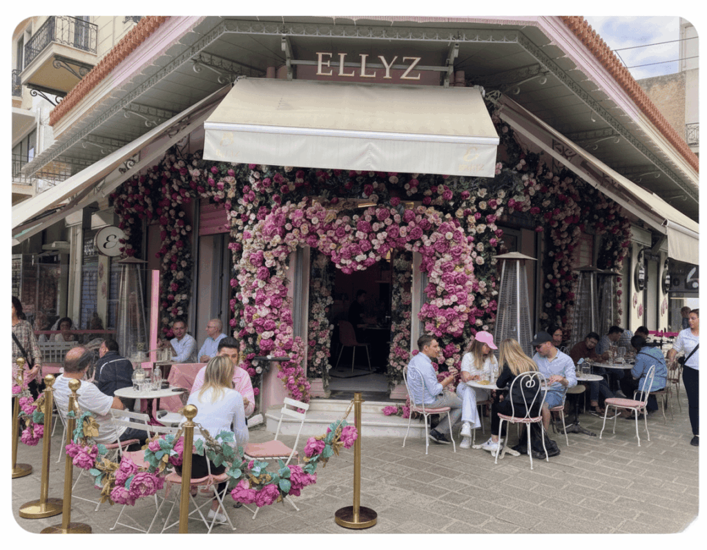 Ein üppig mit pinken und lila Rosen bedecktes Café Ellyz in Athen, dessen Fassade wie ein blühender Garten wirkt, während Gäste draußen an kleinen Tischen die lebendige Atmosphäre genießen.