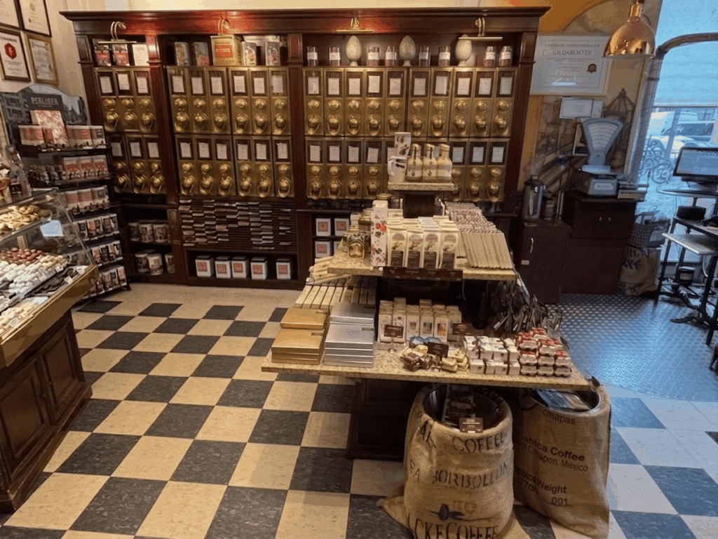 In diesem nostalgisch eingerichteten Fachgeschäft präsentieren sich edler Kaffee und feine Pralinen vor einer imposanten Wand aus goldenen Messingschütten und einem klassischen Schachbrettboden (Bild KI-optimiert).