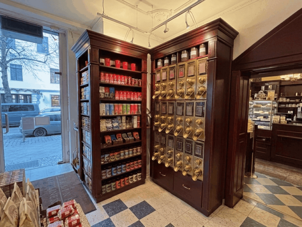 In einem eleganten, dunkelbraunen Holzregal werden zahlreiche Kaffeesorten präsentiert, wobei eine Seite mit bunten Packungen gefüllt ist und die andere über glänzende, goldfarbene Schütten zur Entnahme loser Bohnen verfügt (Bild KI-optimiert).