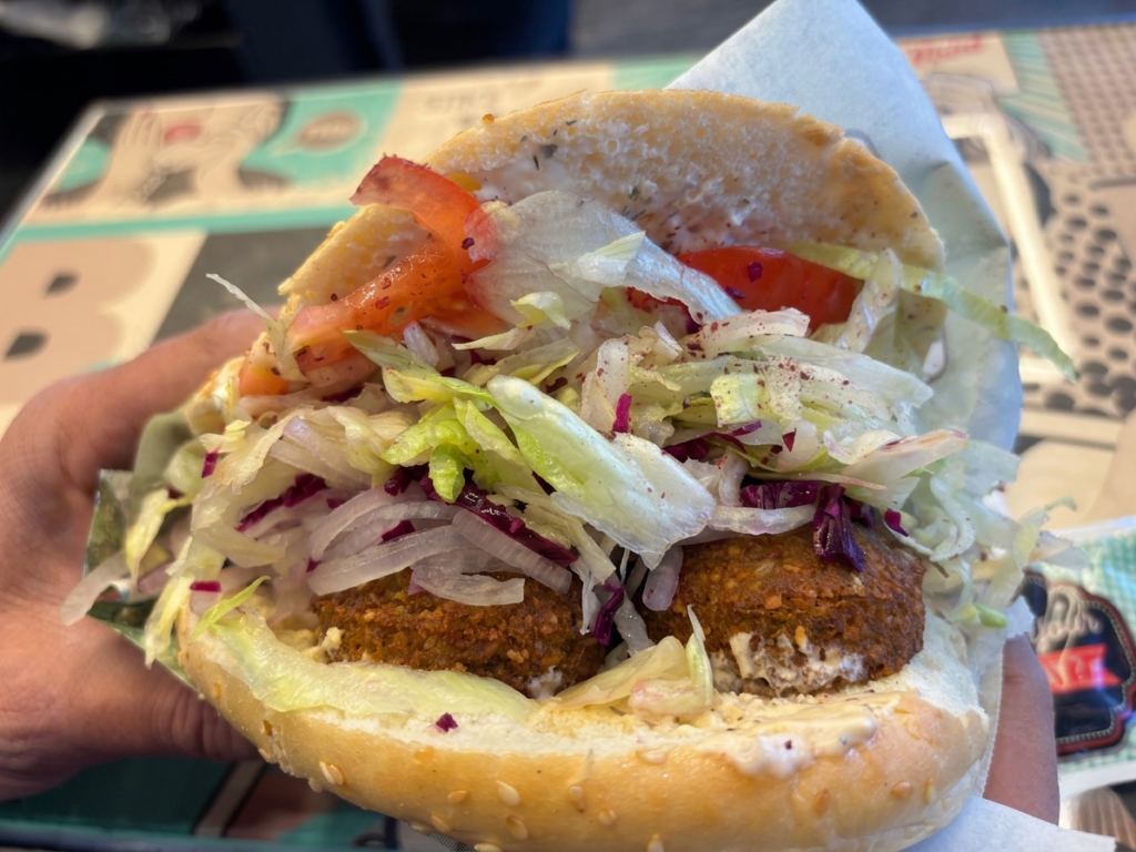 Die Mitarbeiter von ZADDY'S belegen das geöffnete Fladenbrot sorgfältig mit frischem Salat, Tomaten und Rotkohl, während die Kunden geduldig auf den leckeren Falafel warten. 
