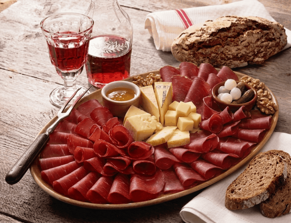 In Graubünden wird das Beisammensein oft mit einer herzhaften „Marenda“ zelebriert, bei der luftgetrocknetes Bündnerfleisch, würziger Bergkäse und regionales Holzofenbrot auf einer Platte kunstvoll angerichtet werden. Diese kulinarische Tradition verbindet handwerkliche Qualität mit purer Geselligkeit und wird idealerweise durch einen kräftigen Wein aus der Region ergänzt.