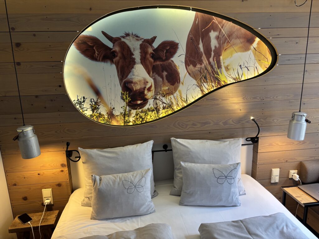Hotelbett in einem Holzverkleideten Zimmer mit einladenden Kissen, zwei Milchkannen-Lampen und einem hinterleuchteten Wandbild einer bayerischen Kuh.