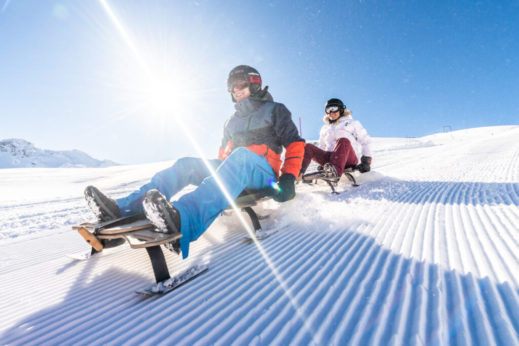 Zwei Personen fahren auf Holzschlitten eine präparierte, verschneite Skipiste bei sonnigem Wetter hinunter.