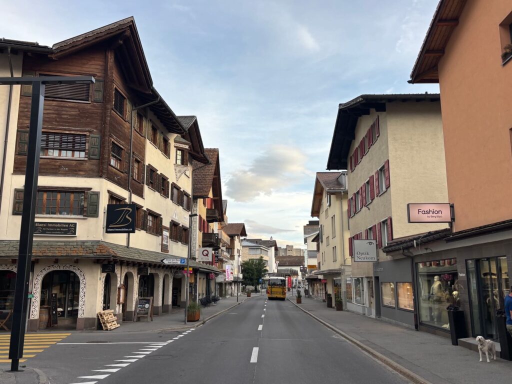 Blick durch die verwaiste Hauptstraße von Lenzerheide mit Bündner Häusern, geschlossenen Geschäften, Gehwegen und kaum Verkehr bei bewölktem Himmel.