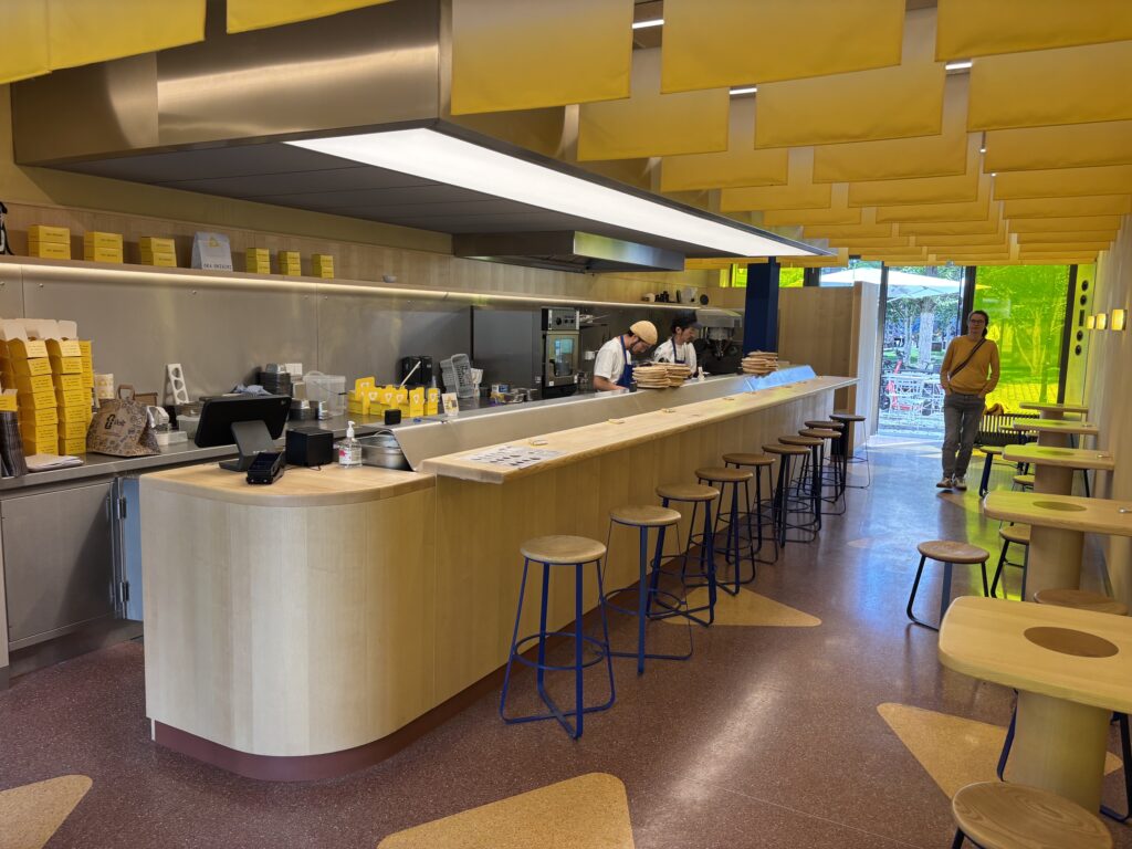 Moderne, puristisch gestaltete Gastro-Bar von OKA Berlin mit hellem Holztresen, blauen Hockern, terrakottafarbenem Boden und gelben Stoffbahnen an der Decke.